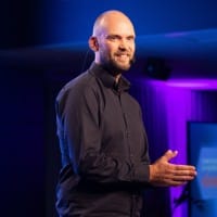 Kuva hymyilevästä miehestä pitämässä puhetta. Tausta violetin sininen.